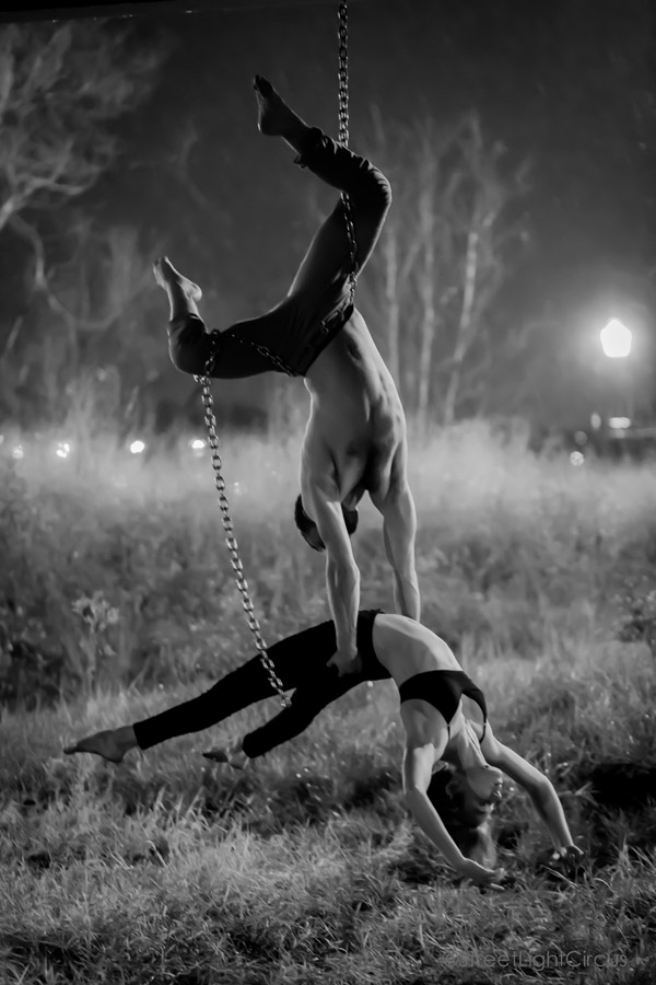 A man suspended upside-down by a chain on his waist using only his hands to hold a woman who leans back parallel to the ground.