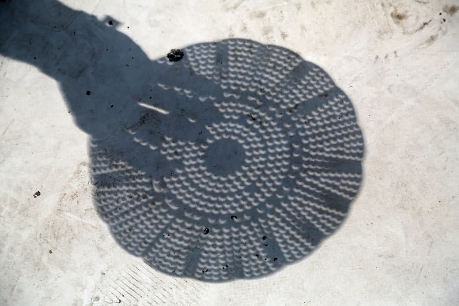 Shadow of hand holding colander projected on ground. The holes of the colander appear crescent-shaped because of the eclipse.