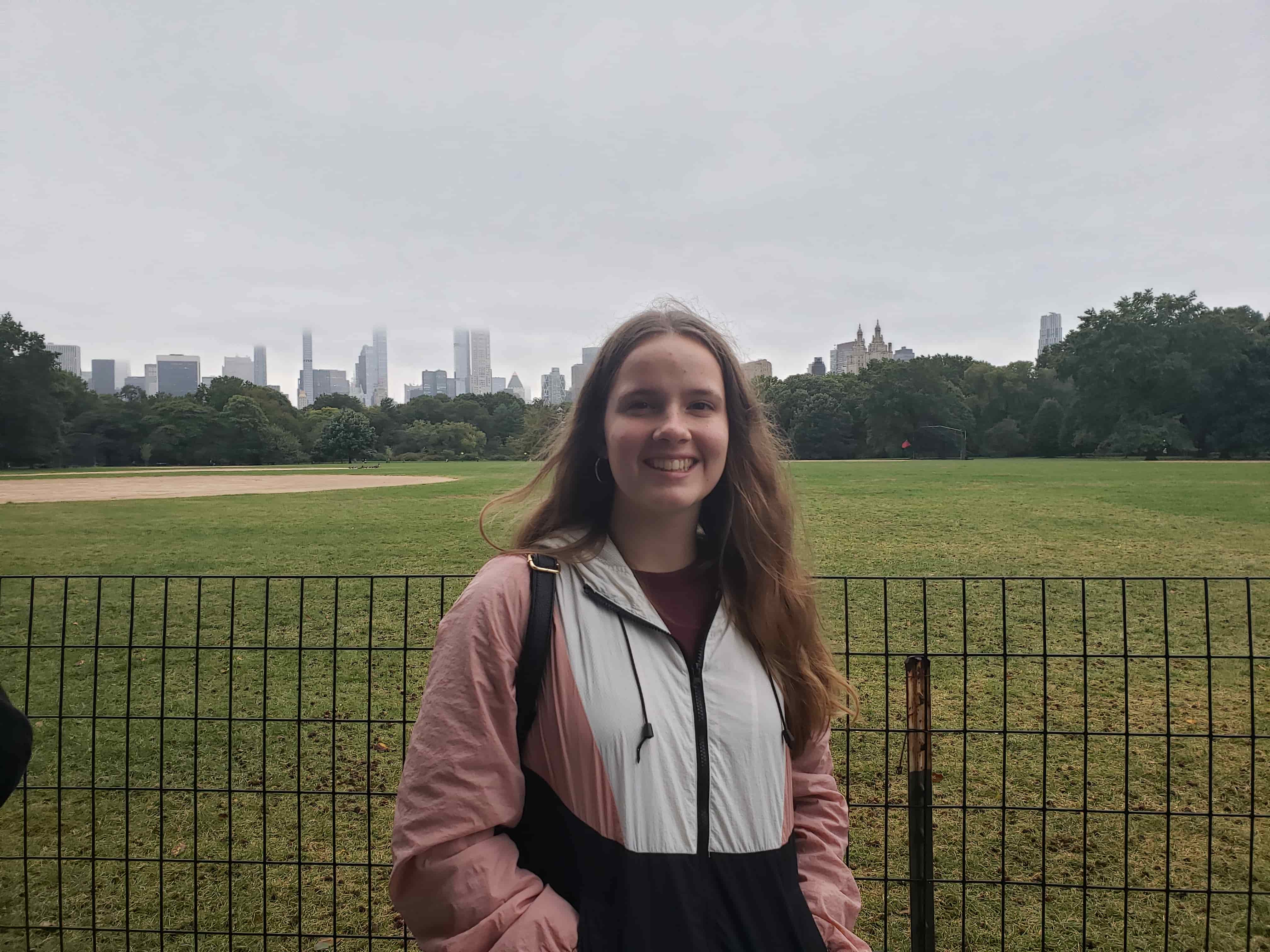 Photo of Melia standing in front of Central Park NYC wearing a pink and white windbreaker. She has long dirty-blonde hair and is smiling at the camera.