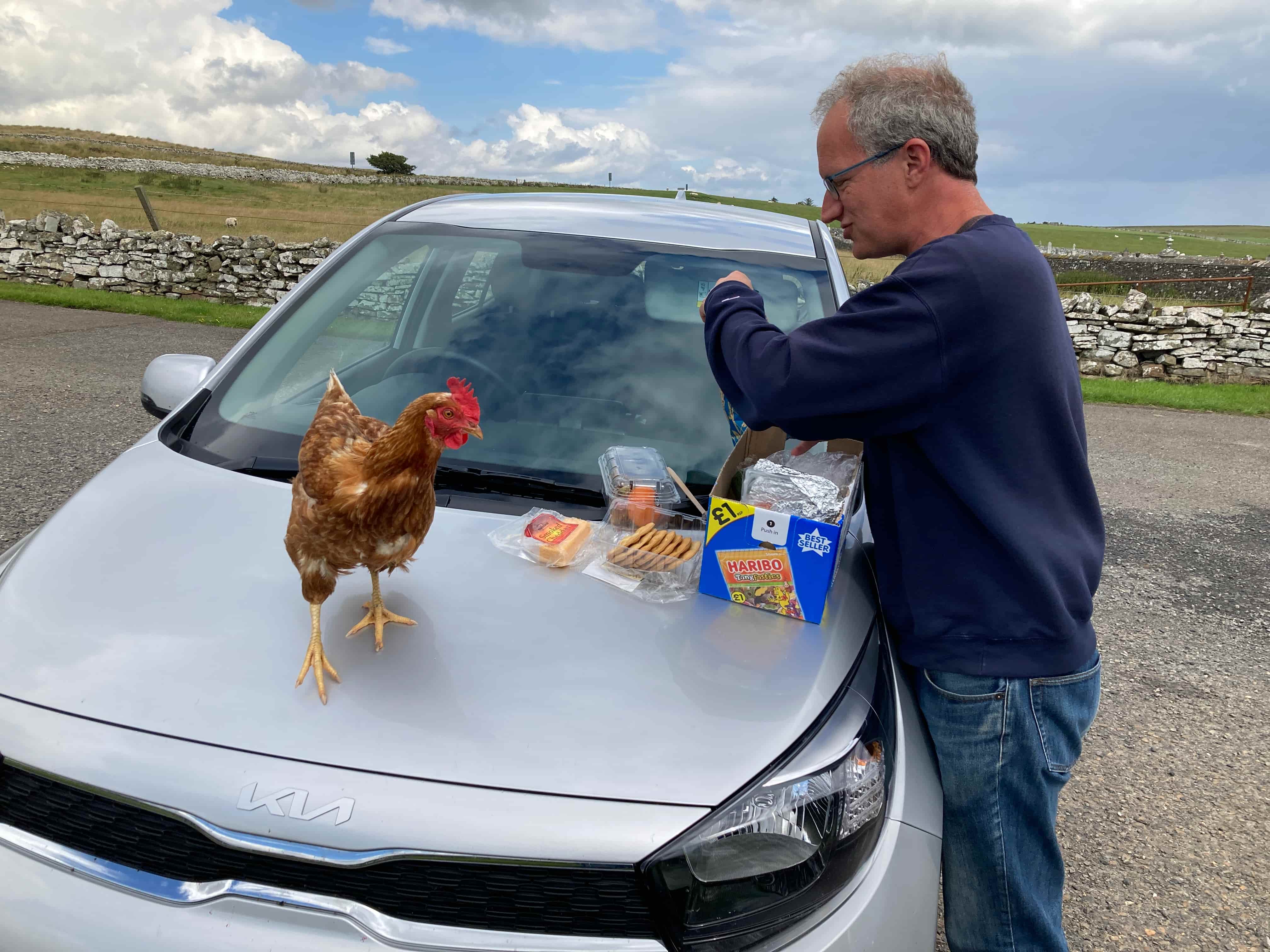 A photo of Prof Adcock standing next to a silver car. On the cars hood are a pile of snacks and a chicken