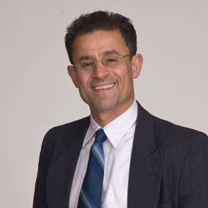 Prof Nimer wearing a white shirt with a blue tie and a black blazer smiling at the camera