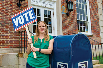 Elizabeth Daigneau stands in front of a blue mail collection bin holding a vote sign