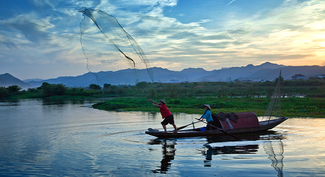 two men throw a fishing net across the water from a small boat with beautiful mountains in distance at sunrise