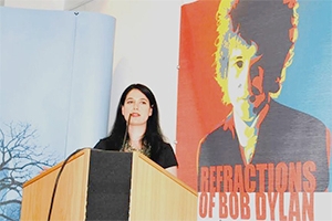A dark-haired Caucasian woman stands at a podium in front of a bright Bob Dylan poster. 