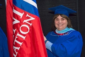 Pat Oltmann posing in her cap and gown next to an alumni relations flag.