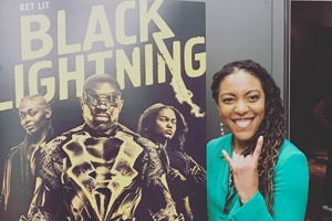 Alumna Anedra Edwards having fun and standing in front of a poster for the CW TV series that she works on, Black Lightning.