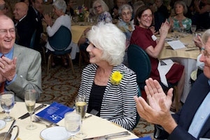 White-haired woman with a flower in her lapel sits at a table as others applaud