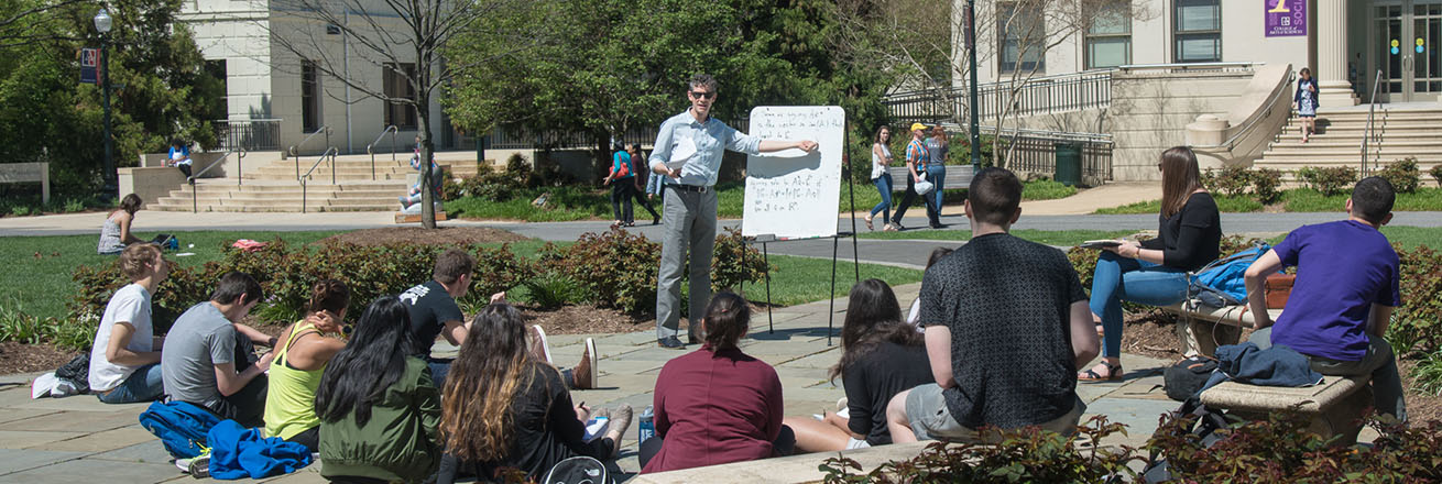 A professor conducts a class discussion outside on the quad on a beautiful sunny day.