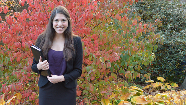 young woman dressed in black skirt suit and purple blouse standing in front of autumn leaves