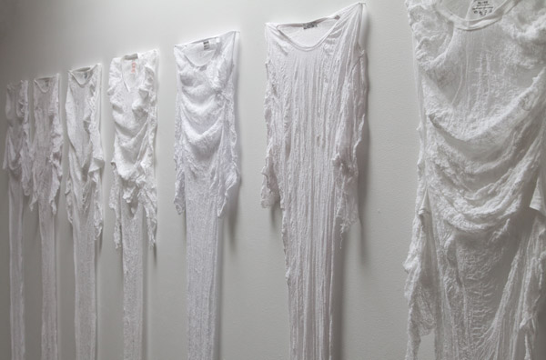 Seven shrunken t-shirts are pinned to the wall: warped, wrinkled, delicate.