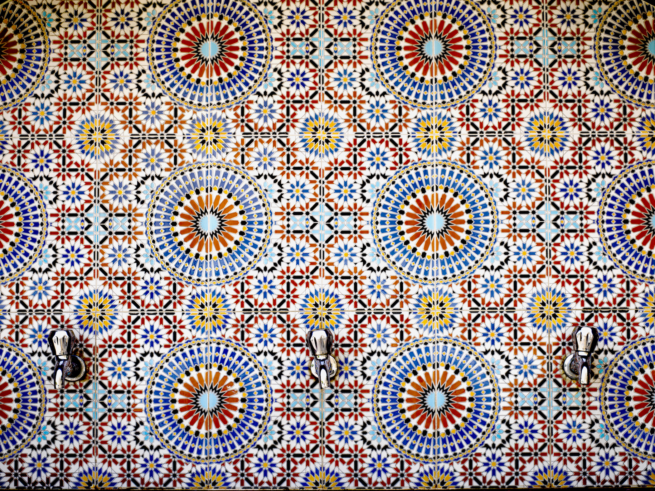 Geometric tiled wall in orange, blue, and yellow with three faucets at the bottom