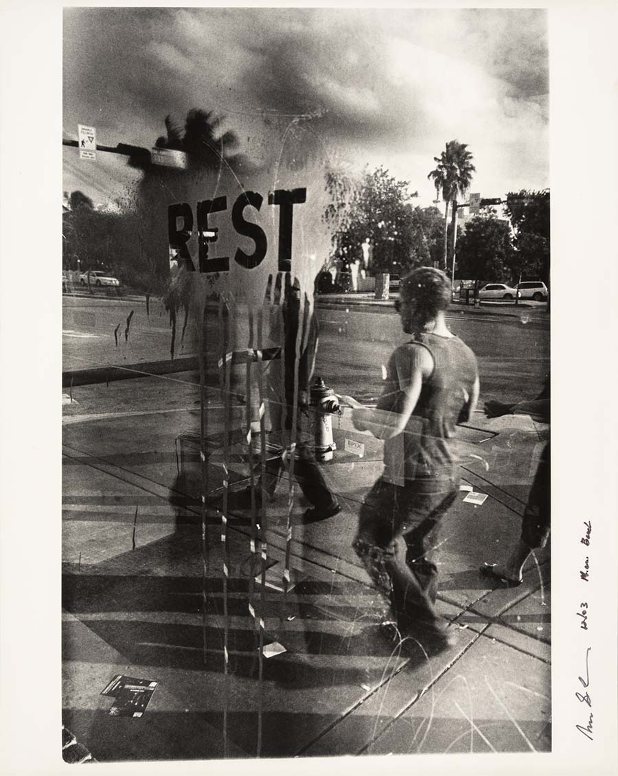 Norman S. Carr, Rest—Miami Beach, Florida, 2003. Black-and-white photo of people walking on a street with 'REST' spraypainted on a window