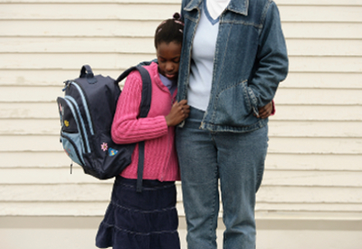 A child holding a backpack clutches a parent's side,
