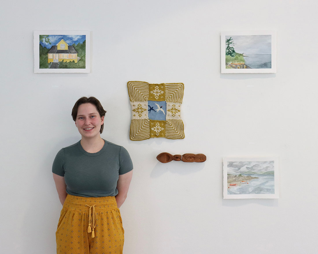Hannah Sjovold with artwork: square textile, carved wood spoon, and paintings of buildings and sea