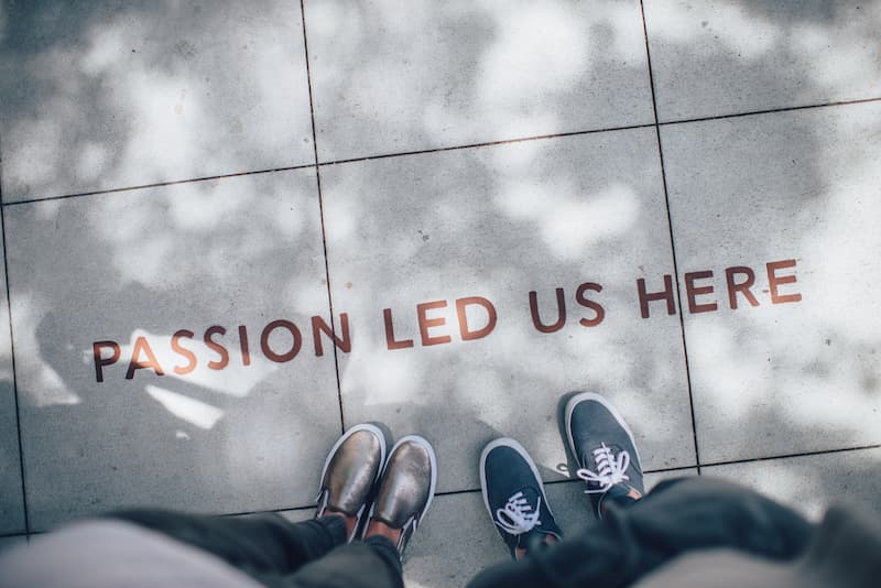 Two people standing on gray tile with Passion Led Us Here; credit: Ian Schneider, Unsplash.