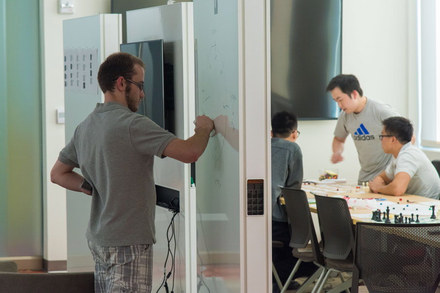 A student writes on a whiteboard wall; on the opposite side, three people lean over a table in conversation