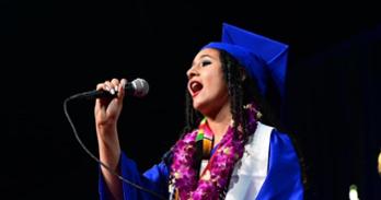 Aisley Wallace Harper, dressed in graduation regalia, sings the national anthem at the May 2023 College of Arts and Sciences graduation ceremony