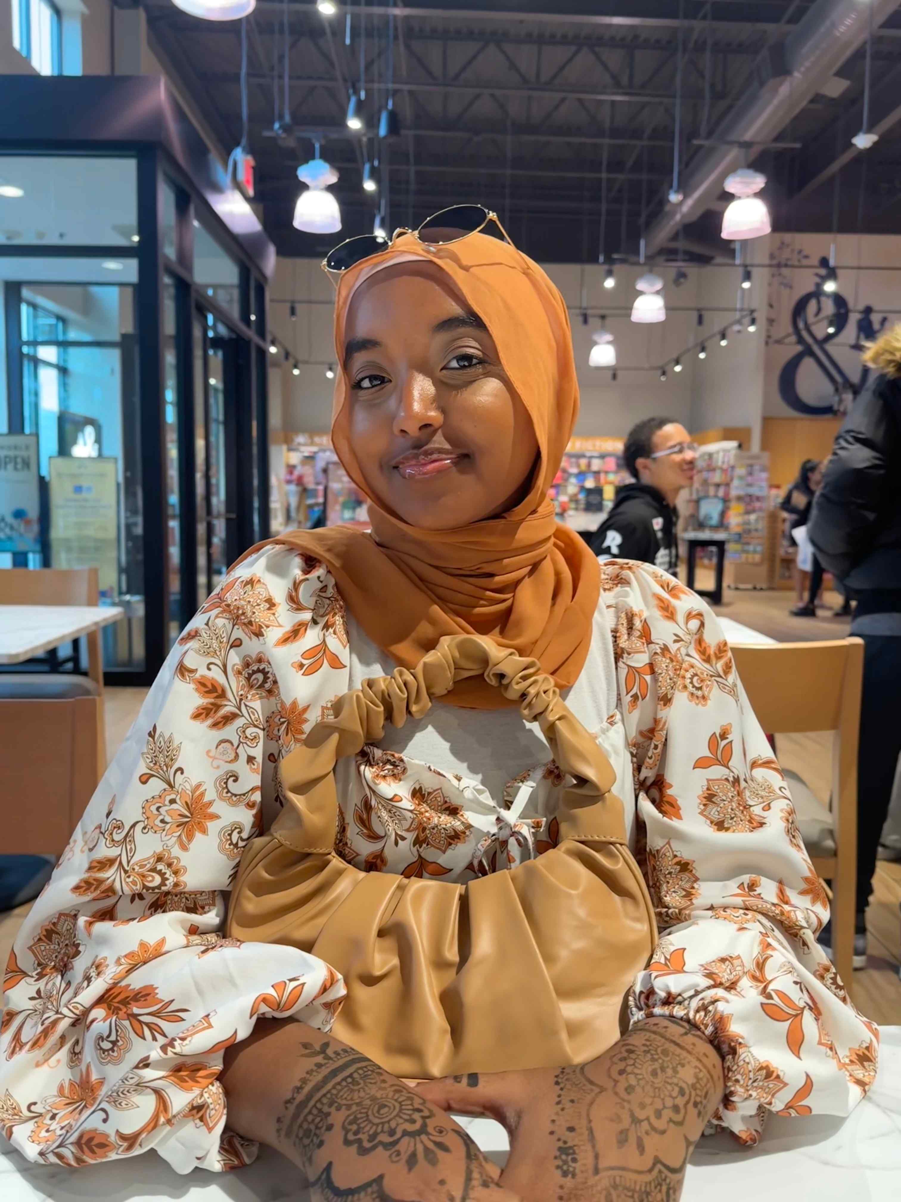 A photo of Aseel wearing an orange head scarf with sunglasses on top and a floral shirt sitting at a restaurant.