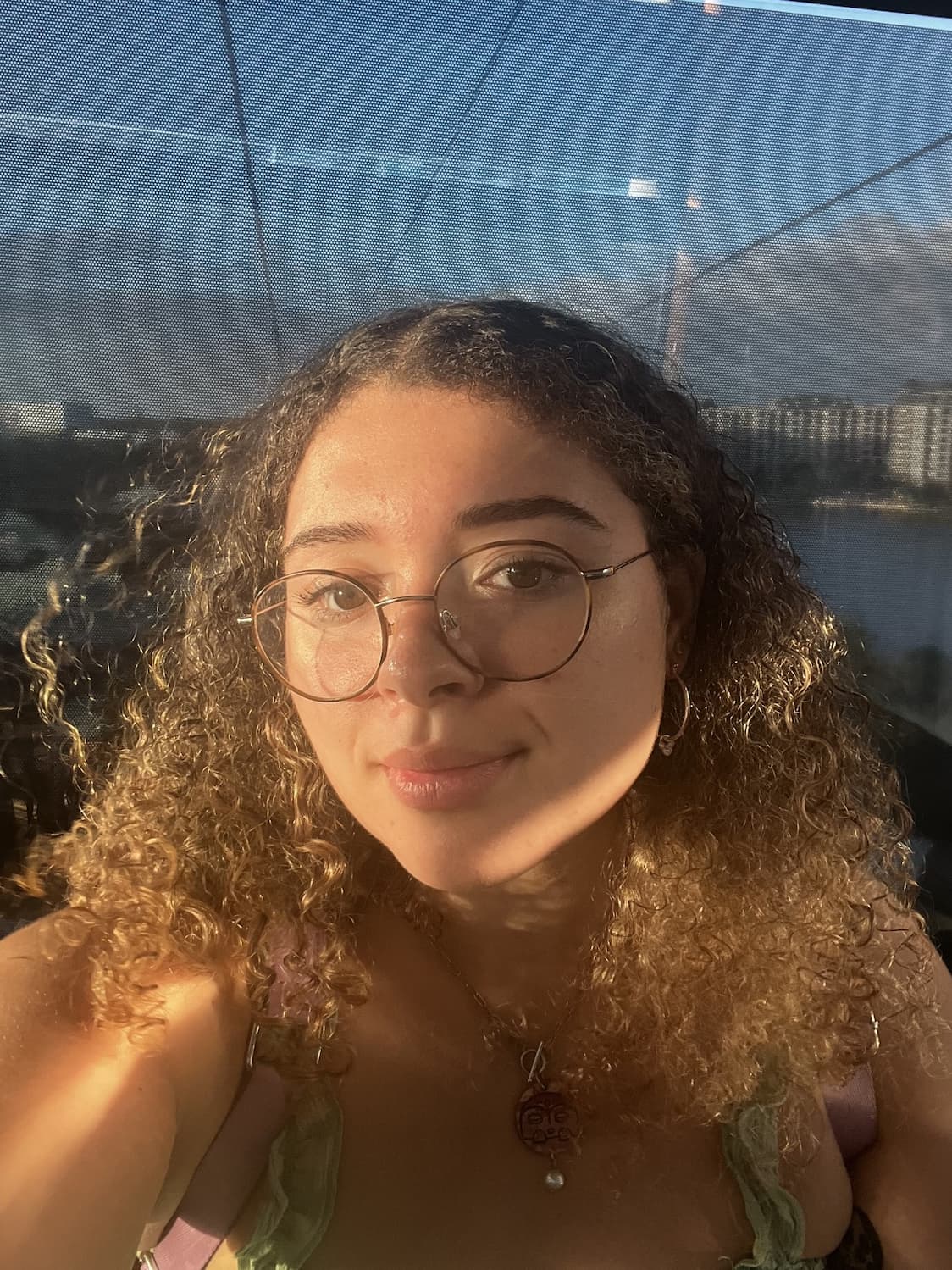 Cheyenne, a young person with medium-dark skin,  glasses and curly brown hair in front of a city and a body of water.