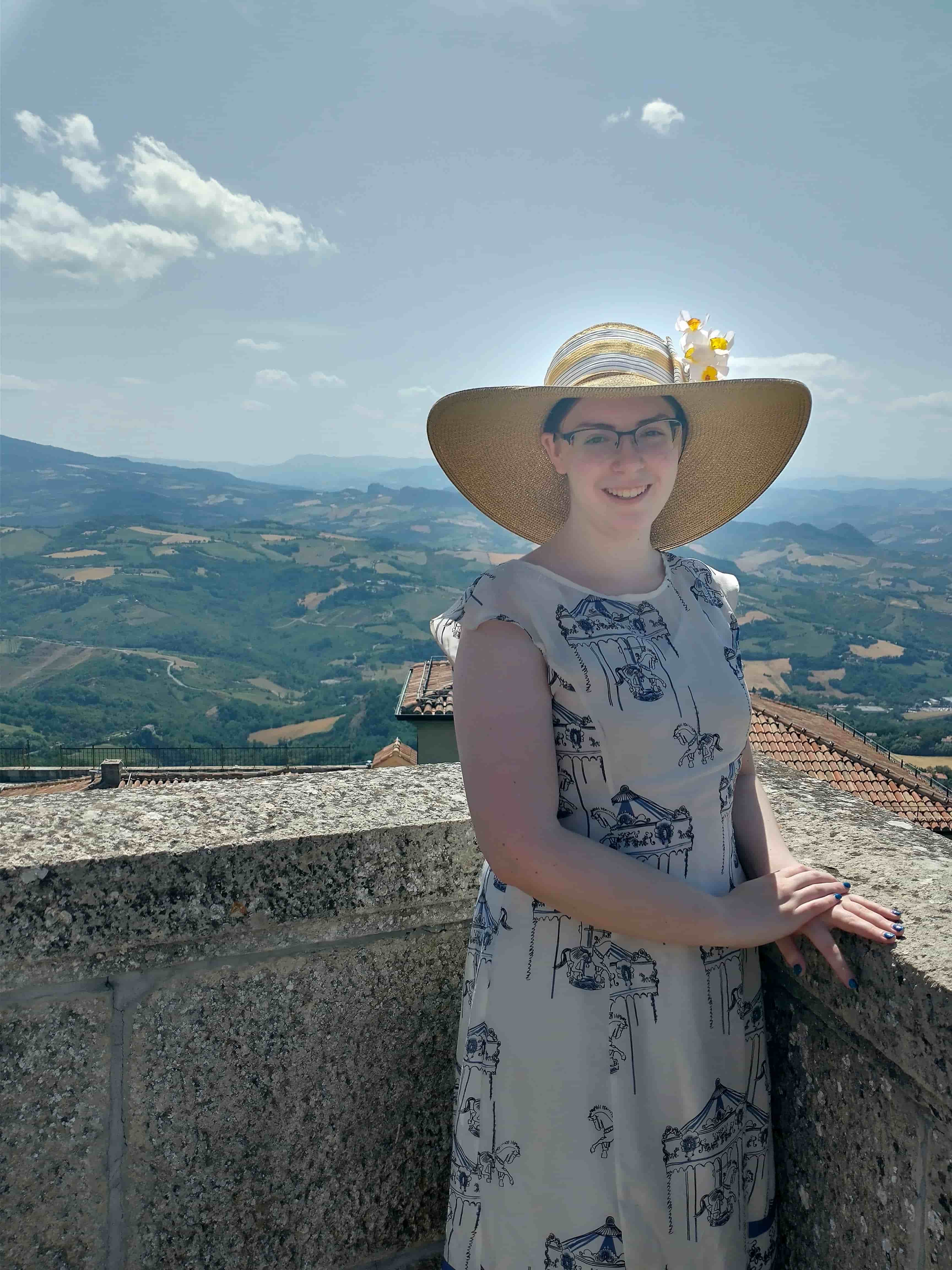 A photo of Abby wearing a sundress and a large straw hat standing in front of a background of hills and fields
