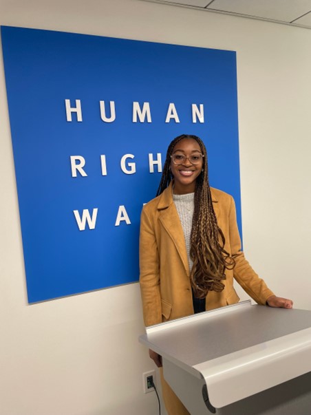 Denia is pictured in a tan jacket and a gray sweater behind a silver podium. Behind Denia is a blue sign that reads “Human Rights Watch” in white text. 