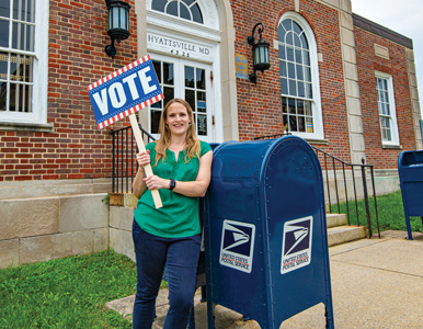 Elizabeth Daigneau stands in front of a blue mail collection bin holding a vote sign