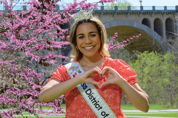 Jude Mabone, wearing a sash and tiara, makes a heart with her hands
