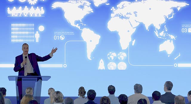 man giving presentation to group of people with world map infographic displayed behind him