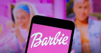A phone screen shows the world "Barbie" and the characters Barbie and Ken are in the background