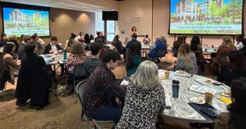 a group of people at round tables looks at someone speaking at a podium during a Preserving the Public Service Pipeline powerpoint slideshow