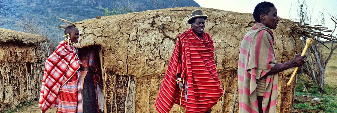 three members of the Maasai dressed in their distinctive bright clothing in front of their mud huts in Kenya