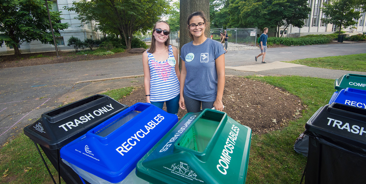Two AU students smile while standing behind some of the university's trash, recycling, and composting receptacles.