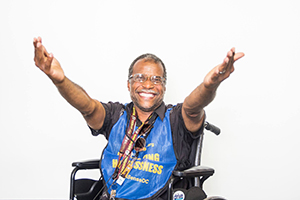 A man in a Street Sense smock in a wheelchair smiling and raising his open arms