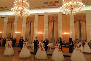Women in white dresses with red sashes stand in an elegant ballroom with their partners.