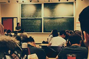 Students facing a chalkboard with equations on it and a professor standing next to it, teaching